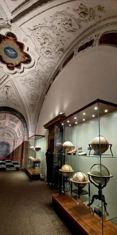 Corridor inside the Strahov library featuring ornate stucco ceilings and a glass display of historical globes and scientific instruments, illuminated by soft museum lighting.