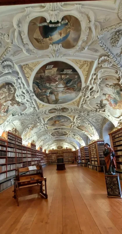 Interior of the Strahov library in Prague, featuring long wooden floors, walls lined with ancient books, and an ornate Baroque ceiling decorated with elaborate frescoes and stucco details.