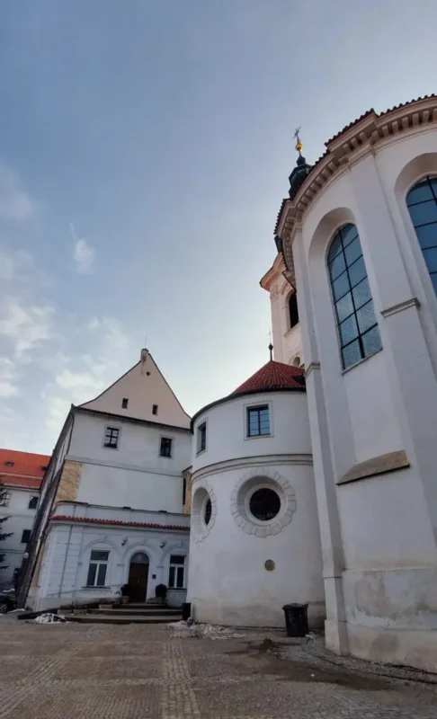 Courtyard near the Strahov library in Prague, showing historic white monastery buildings with rounded towers, arched windows, and red-tiled roofs under a clear sky.