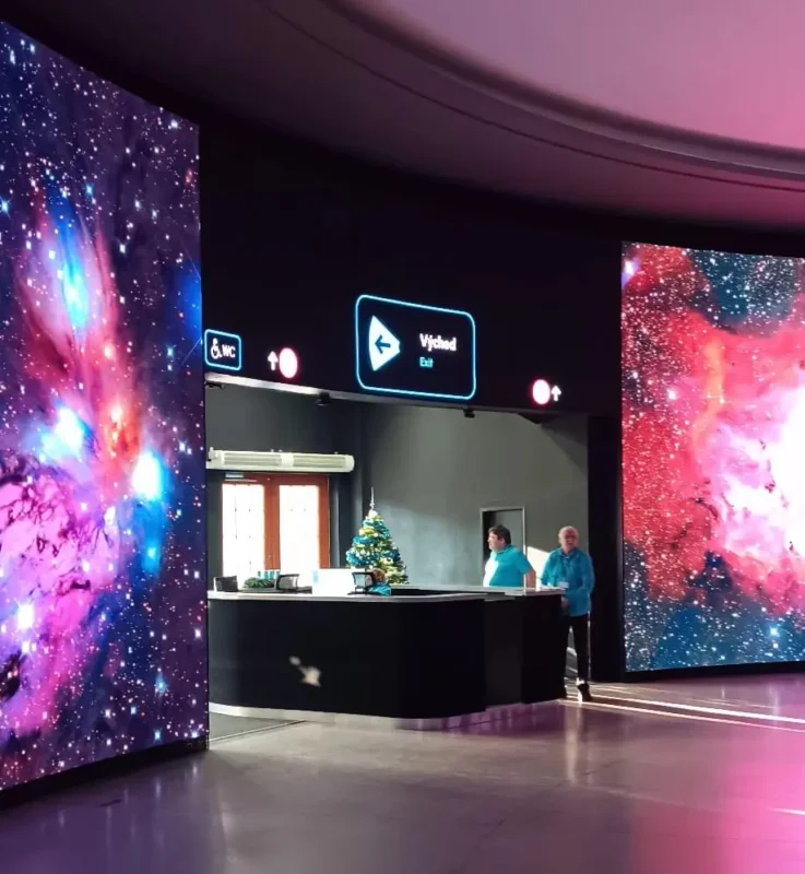 Interior of the Prague Planetarium featuring large LED walls displaying colorful galaxy imagery, a modern reception desk, and illuminated exit signage in a dimly lit, futuristic space.