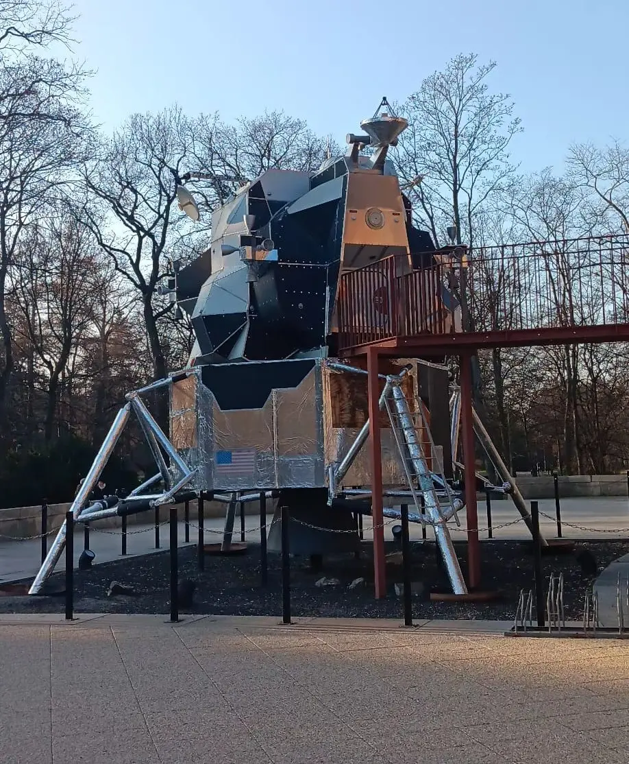 Outdoor space probe exhibit near the Prague Planetarium, displayed on metal supports beside the circular planetarium building, with trees and clear blue sky in the background.