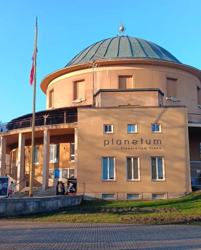 Exterior view of the Prague Planetarium with its circular building and large dome, photographed on a clear day, with the Czech flag standing in front of the entrance.