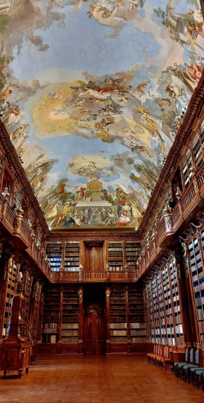 Interior of the Strahov library’s Baroque Theological Hall, featuring towering wooden bookshelves, ornate carved details, and a large ceiling fresco depicting heavenly scenes.