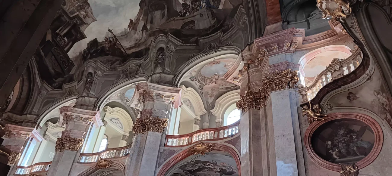 **Alt text (EN):**
*Baroque interior of St. Nicholas Church in Prague, featuring curved arches, marble columns with gilded capitals, ornate balconies, and ceiling frescoes illuminated by natural light from tall windows.*