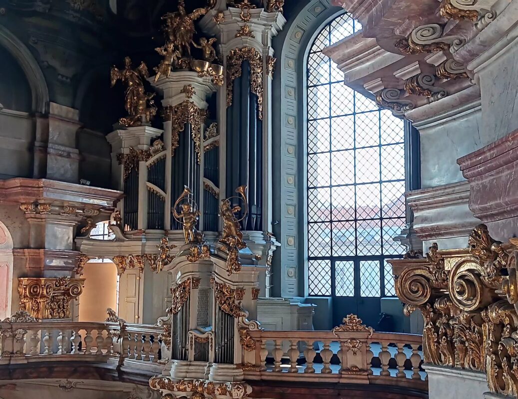Ornate Baroque church interior featuring a grand pipe organ adorned with gilded angel sculptures, decorative columns, and a large arched window letting in natural light.”