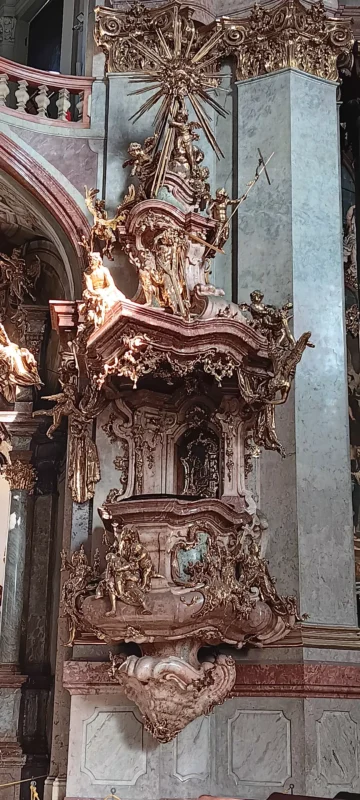 Highly ornate Baroque pulpit inside a church, richly decorated with gilded sculptures of angels, saints, and dramatic figures, topped by a radiant sunburst and surrounded by marble columns