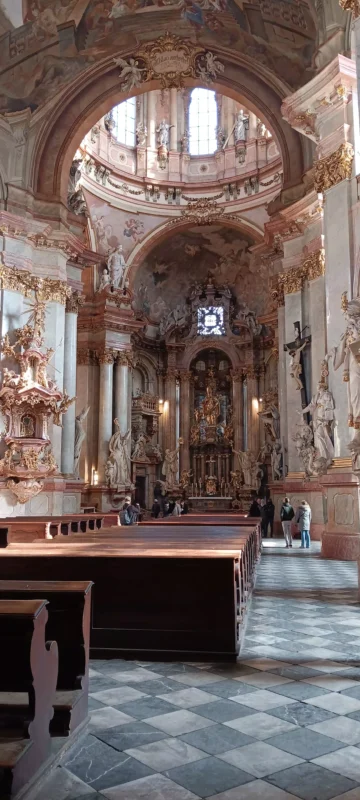 **Alt text (EN):**
*Interior of St. Nicholas Church in Prague, showing the grand Baroque nave with marble columns, gilded sculptures, frescoed arches, wooden pews, and the main altar beneath the high dome, with visitors inside.*